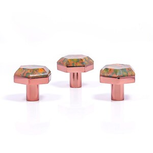 Op de afbeelding: Drie decoratieve kastknoppen met een geometrisch ontwerp. De knoppen hebben een koperkleurige basis en een facetgeslepen bovenkant met een mix van groene, oranje en gele kleuren. Ideaal voor meubels en woondecoratie.
