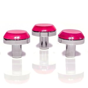 Puede incluir: Tres pomos decorativos para armarios. Cada pomo presenta un diseño bicolor rosa y plateado. La parte superior es de color rosa brillante, con un acento rosa más claro. La base es de metal plateado brillante. Los pomos están dispuestos sobre una superficie blanca.