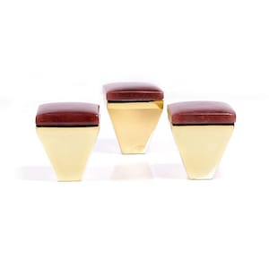 Könnte beinhalten: Drei dekorative Boxen mit geometrischem Design. Jede Box hat eine quadratische, dunkelrote Oberseite und eine goldfarbene, trapezförmige Basis. Die Boxen sind auf weißem Hintergrund angeordnet.