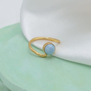 Könnte beinhalten: Ein goldfarbener Ring mit einem hellblauen Edelstein. Der Ring hat ein schlichtes, offenes Design, wobei der Edelstein in einer kreisförmigen Fassung gefasst ist. Der Ring wird auf weißem Stoff und einer hellgrünen Oberfläche präsentiert.