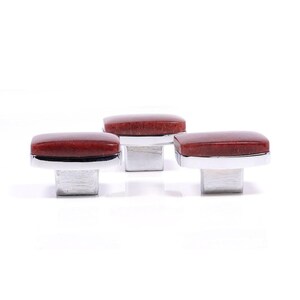 Puede incluir: Tres tiradores decorativos para armarios. Cada tirador presenta una piedra rectangular de color marrón rojizo sobre una base de metal plateado. Los tiradores son cuadrados y tienen un acabado pulido, aptos para muebles o decoración del hogar.