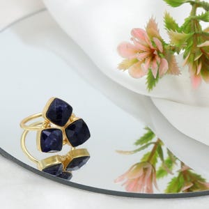 Może przedstawiać: Złoty pierścionek z trzema ciemnoniebieskimi kwadratowymi kamieniami. Pierścionek jest umieszczony na odblaskowej powierzchni, z rozmytym tłem z białej tkaniny i różowych kwiatów.