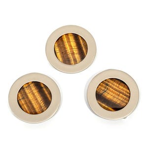May include: Three circular decorative objects. Each has a light-colored metal ring surrounding a polished, round stone with brown and gold tiger's eye patterns. The objects are arranged on a white background.