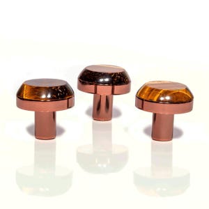 May include: Three mushroom-shaped cabinet knobs with a copper finish and tiger's eye stone tops. Each knob has a round, polished stone set into a copper base. The knobs are displayed on a white surface, reflecting the light.