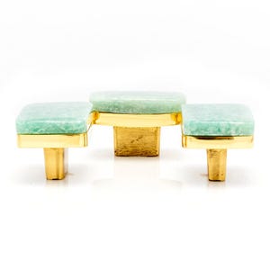 May include: Three cabinet knobs with square, light green stone tops and gold-colored bases. The knobs are arranged in a staggered pattern, with the center knob slightly higher than the two side knobs.