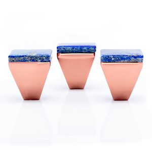 Op de afbeelding: Drie koperkleurige, geometrisch gevormde decoratieve objecten met een vierkante, blauwe stenen bovenkant. De objecten worden gepresenteerd op een wit oppervlak. De koperkleurige basis heeft een driehoekige vorm.