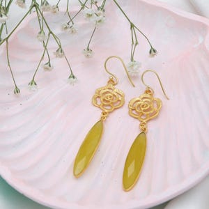 May include: Gold-coloured earrings featuring a floral design and light green, teardrop-shaped gemstones. The earrings are presented on a pink, shell-shaped dish, with white flowers in the background.