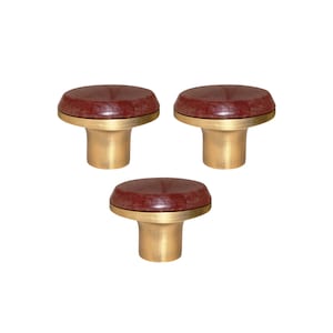 May include: Three cabinet knobs with a dark red, circular top and a brass-coloured base. The knobs have a simple, elegant design, suitable for drawers or cabinets. The knobs are shown against a white background.