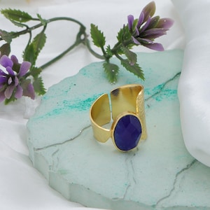 Op de afbeelding: Een goudkleurige ring met een grote, ovale, diepblauwe edelsteen. De ring heeft een getextureerde band en een open ontwerp. De edelsteen is facetgeslepen en vangt het licht op. De ring wordt gepresenteerd op een lichtblauwe, getextureerde ondergrond.