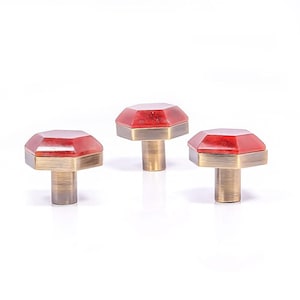 May include: Three cabinet knobs with faceted red tops and brass-coloured bases. The knobs have an octagonal shape. They are set against a white background. These knobs are suitable for furniture or home decor.
