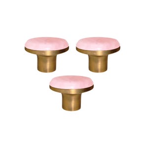 Pode incluir: Três puxadores de armário com um topo arredondado rosa claro e uma base de cor latão. Os puxadores têm um design simples e elegante, adequado para gavetas ou armários. Os puxadores estão em um fundo branco.