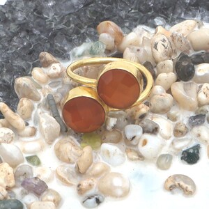 Puede incluir: Un anillo dorado con dos gemas redondas, facetadas, de color rojo anaranjado. El anillo se encuentra sobre un fondo de pequeños guijarros y piedras multicolores, creando una estética natural y terrosa. Las gemas son el foco principal de la joya.