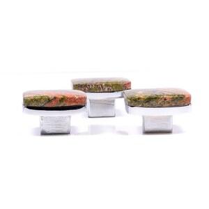 May include: Three cabinet knobs with rectangular unakite stone tops in shades of red, green, and brown. The knobs have a silver-coloured metal base and a square post. The knobs are set against a white background.