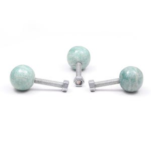 Può includere: Tre pomelli decorativi per mobili con finitura verde chiaro, simile al marmo. Ogni pomello ha una forma sferica attaccata a un bullone e un dado argentati. I pomelli sono disposti su uno sfondo bianco.