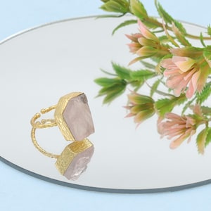 Puede incluir: Un anillo dorado con una gema rectangular grande de color rosa claro. El anillo está sobre una superficie reflectante, con un arreglo floral en el fondo. La gema es cuarzo rosa.