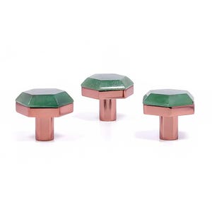 Könnte beinhalten: Drei dekorative Schrankknöpfe. Jeder Knopf hat eine facettierte, smaragdgrüne Edelsteinspitze und einen kupferfarbenen Sockel. Die Knöpfe sind vor einem weißen Hintergrund angeordnet.