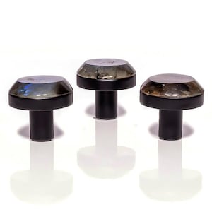 Op de afbeelding: Drie decoratieve kastknoppen met een paddenstoelachtig ontwerp. Elke knop heeft een zwarte basis en steel, bekroond met een metallic, iriserende dop. De doppen reflecteren verschillende kleuren en geven het ontwerp een vleugje elegantie.