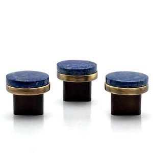 Può includere: Tre pomelli decorativi per mobili. Ogni pomello presenta una parte superiore rotonda in pietra blu scuro con una fascia metallica color oro, posta su una base quadrata marrone scuro. I pomelli sono disposti in fila su sfondo bianco.