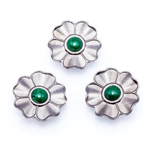 May include: Three silver-coloured flower-shaped buttons with a green, circular centre. The buttons have a detailed petal design and are set against a white background. These decorative buttons could be used for sewing or crafts.