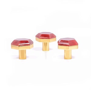 May include: Three cabinet knobs with a red, faceted, gem-like top and a gold-coloured base and stem. The knobs are displayed against a white background, showcasing their jewel-like appearance. The knobs are suitable for cabinets.
