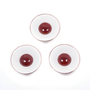 May include: Three white and red decorative coasters. Each coaster has a white outer ring with concentric gray lines, a red center, and a thin copper-colored rim. The coasters are arranged in a triangular pattern against a white background.