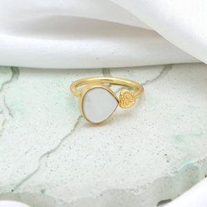 May include: Gold-tone ring featuring a pear-shaped white stone and a small heart-shaped accent. The ring is set on a light-colored surface with a white fabric in the background. The ring's design is simple and elegant.