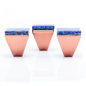 May include: Three copper-colored, geometric-shaped decorative objects with a square, blue stone top. The objects are displayed on a white surface, with a reflection visible below. The design is modern and minimalist.