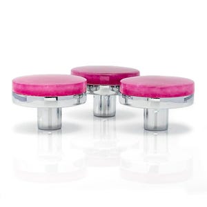 May include: Three cabinet knobs with a vibrant pink, circular top and a polished, silver-coloured base. The knobs are grouped together, with a reflective surface beneath them.