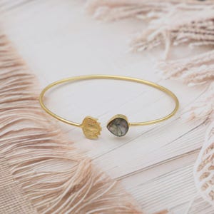 May include: Gold-coloured bangle bracelet with a leaf-shaped detail and a teardrop-shaped gemstone. The open-ended bracelet features a leaf and a gemstone. The gemstone displays a mix of grey and green tones.
