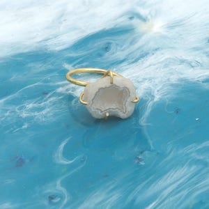 Op de afbeelding: Een goudkleurige ring met een lichtgekleurde, geode-achtige steen. De steen heeft een natuurlijke, onregelmatige vorm met een contrasterend binnenpatroon. De ring staat tegen een blauw-witte achtergrond.