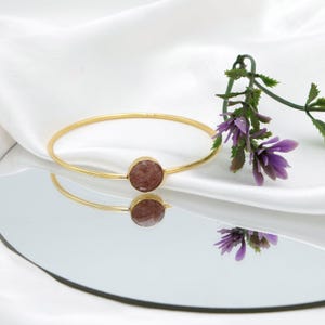 Può includere: Un bracciale a cerchio dorato con una pietra rotonda marrone-rossastra al centro. Il bracciale è esposto su una superficie riflettente con un tessuto bianco e fiori viola sullo sfondo. Il design del bracciale è semplice ed elegante.