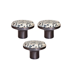 Op de afbeelding: Drie decoratieve kastknoppen. Elke knop heeft een ronde, gespikkelde, gebroken witte en zwarte bovenkant, geplaatst op een donkerbruine, cilindrische basis. De knoppen zijn in een driehoekspatroon tegen een witte achtergrond geplaatst.