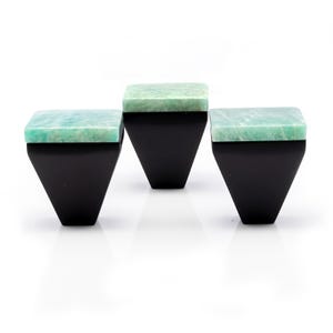Könnte beinhalten: Drei dekorative Schrankknöpfe. Jeder Knopf hat eine quadratische, hellgrüne Steinplatte und einen schwarzen, trapezförmigen Sockel. Die Knöpfe sind vor einem weißen Hintergrund angeordnet.