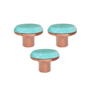 May include: Three decorative cabinet knobs. Each knob features a round, turquoise-colored top with a copper-colored base. The knobs are arranged in a triangular pattern against a white background.