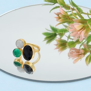 May include: A gold-toned ring with a cluster of gemstones. The ring features a large oval black stone, surrounded by smaller green, white, and black stones. The ring is displayed on a reflective surface with floral accents.