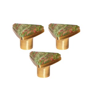 Può includere: Tre pomelli per mobili con una parte superiore triangolare in pietra screziata verde e rossa. I pomelli hanno una base e finiture in metallo color oro, su sfondo bianco.