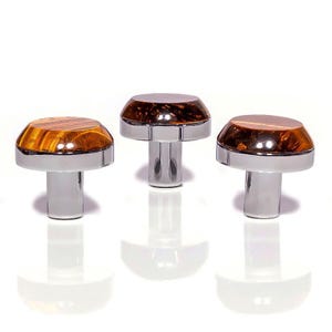 Può includere: Tre pomelli decorativi per mobili a forma di fungo. Ogni pomello presenta una base cromata lucida e una parte superiore a cupola con un motivo ambrato o marrone traslucido. Presentati su sfondo bianco.