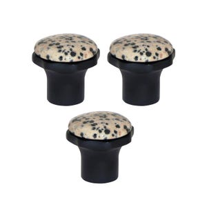 Op de afbeelding: Drie zwarte kastknoppen met gespikkelde beige en zwarte bovenkanten. De knoppen hebben een paddenstoelvorm, met een afgeronde bovenkant en een uitlopende basis. De knoppen zijn waarschijnlijk gemaakt van steen of een vergelijkbaar materiaal.