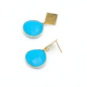 Może przedstawiać: Para złotych kolczyków z kwadratową górą i łezkowatym, fasetowanym, jasnoniebieskim kamieniem. Kolczyki są umieszczone na białym tle.