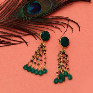 May include: Gold-toned earrings featuring dark green circular studs and dangling green teardrop beads. The earrings have a chain of small purple beads. A peacock feather is in the background.