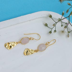 Puede incluir: Un par de pendientes dorados con textura martillada y gemas ovaladas de color rosa pálido. Los pendientes se muestran sobre una bandeja blanca sobre un fondo azul. Los pendientes tienen cierre de gancho.
