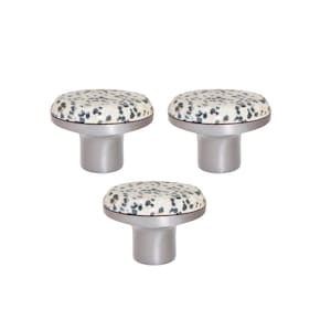 Op de afbeelding: Drie decoratieve kastknoppen. Elke knop heeft een ronde, gespikkelde, gebroken witte bovenkant met grijze en zwarte vlekken, een zilverkleurige metalen rand en een cilindrische zilverkleurige basis. De knoppen zijn in een driehoek gerangschikt.