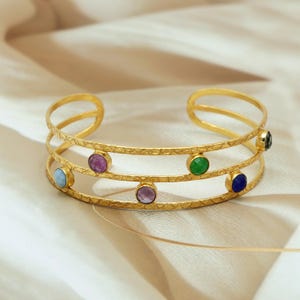 May include: Gold-coloured cuff bracelet with three parallel bands. The bracelet features colourful gemstones, including blue, purple, green, and a dark gemstone. The textured surface adds a unique touch to this jewellery.