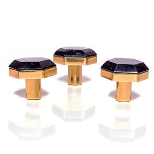 May include: Three cabinet knobs with a dark blue, faceted top and gold-coloured accents. Each knob has a cylindrical gold-coloured base. The knobs are displayed on a white surface.