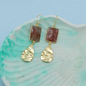 May include: Gold-tone earrings with rectangular, faceted, reddish-brown stones. Below each stone hangs a textured, gold-colored, teardrop-shaped charm. The earrings are displayed on a teal and white marbled surface.