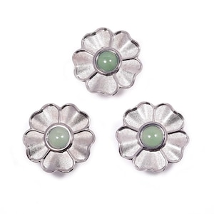 Może przedstawiać: Trzy srebrne elementy biżuterii w kształcie kwiatów z jasnozielonym kamieniem centralnym. Płatki mają teksturowany wygląd. Elementy są ułożone na białym tle.