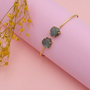 Könnte beinhalten: Ein offenes Armreif-Armband in Goldfarbe mit zwei grau-grünen, unregelmäßig geformten Steinen. Das Armband befindet sich vor einem rosa Hintergrund mit gelben Blumen in der oberen linken Ecke. Die Steine werden von goldfarbenen Zinken gehalten.