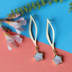 Könnte beinhalten: Ein Paar goldfarbene Ohrringe mit einem blattartigen Design. Jeder Ohrring hat einen sternförmigen Anhänger mit weißem und rosa Farbton. Die Ohrringe werden auf einem blau-rosa Hintergrund mit rosa Blüten und grünen Stielen präsentiert.