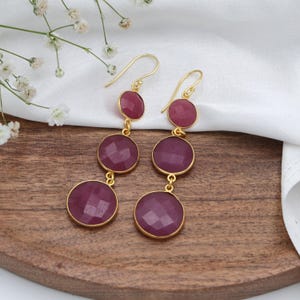May include: Gold-tone drop earrings with three faceted, round, deep pink gemstones. The earrings have a simple hook design. They are displayed on a wooden surface with white fabric and delicate white flowers in the background.