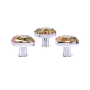 May include: Three cabinet knobs with a faceted, octagonal top made of green and orange speckled stone. The knobs have a silver-coloured metal base and stem. The knobs are displayed against a white background.